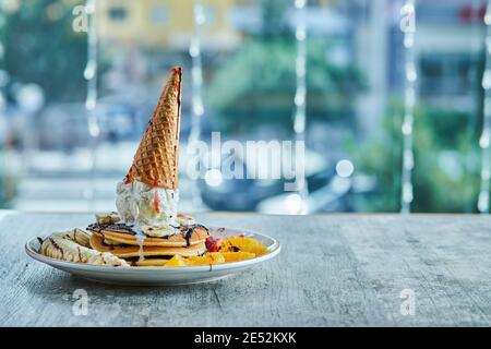 Frittelle con cono gelato, mandarino, fragola, banana e sciroppo di cioccolato nella piastra bianca sullo sfondo di marmo Foto Stock