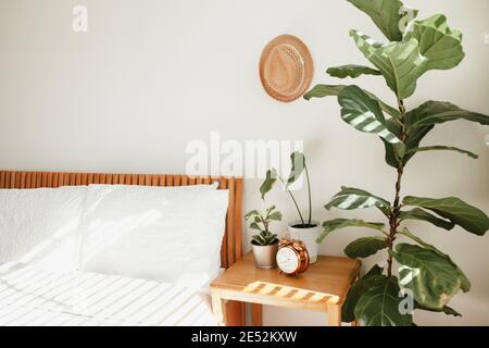 camera da letto bianca con letto in legno arancione, biancheria da letto bianca, comodino e piante vicino al letto, cappello di paglia sulla parete e orologio in rame vintage o Foto Stock