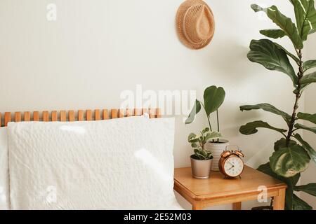 camera da letto bianca con letto in legno arancione, biancheria da letto bianca, comodino e piante vicino al letto, cappello di paglia sulla parete e orologio in rame vintage o Foto Stock