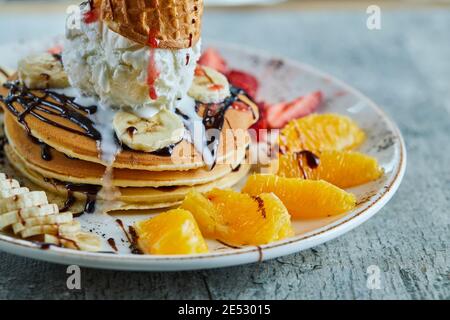 Frittelle con cono gelato, mandarino, fragola, banana e sciroppo di cioccolato nella piastra bianca sullo sfondo di marmo Foto Stock