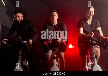 tre uomini impegnati in un simulatore di bicicletta in palestra, allenandosi su una cyclette, isolati in uno spazio fumoso illuminato al neon scuro Foto Stock