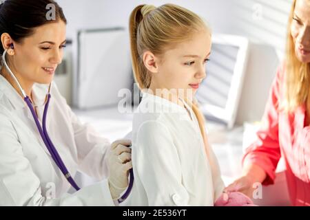 ragazza visiting doctor.female pediatra che controlla i polmoni del piccolo paziente, ascoltando il respiro della ragazza attraverso stetoscopio durante il check-up in clinica o. Foto Stock