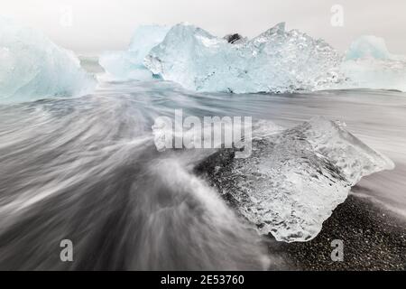 Primo piano di un blocco di ghiaccio che si fonde su una riva nera con acqua che cola e scorre, e iceberg blu chiaro sullo sfondo Foto Stock