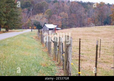 Pali di legno di una recinzione di filo spinato con offset di filo elettrico che conduce ad un vecchio fienile in lontananza in un bel giorno di autunno. Foto Stock