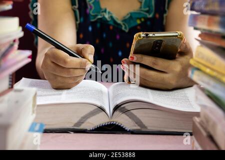 fuoco selettivo su una mano umana femminile che tiene la penna della sfera e telefono cellulare attraverso la pila di libri Foto Stock