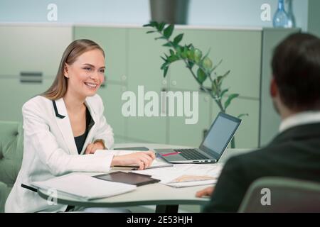 Sorridente agente professionista HR che fa un colloquio di lavoro ad un candidato nel luminoso ufficio moderno. Giovane donna intervistando giovane freelance per lavoro Foto Stock