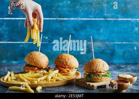 Una mano donna che tiene la patata fritto con tre cheeseburgers, ketchup, maionese su piastra di legno Foto Stock