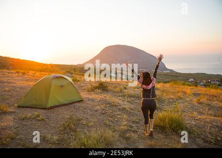 La donna incontra l'alba in montagna, gioisce al sole. Vista panoramica della montagna e del mare dall'alto. Campeggio, attività all'aperto, sport Foto Stock