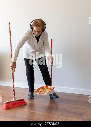 Allegra donna di 90 anni che ascolta la musica in cuffia mentre ampio pavimento con scopa e lavori di casa Foto Stock