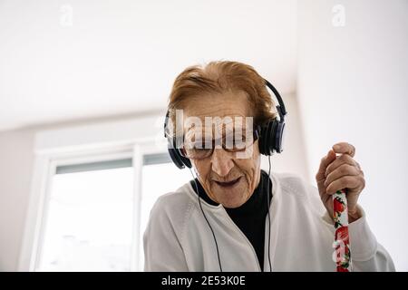 Donna anziana ottimista in cuffia per ascoltare musica e divertirsi canzoni mentre facendo lavoro di casa con scopa Foto Stock