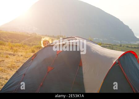 Un bambino allegro si affaccia da dietro una tenda turistica in montagna con una vista panoramica all'alba. Turismo domestico, viaggio estivo attivo, adven famiglia Foto Stock