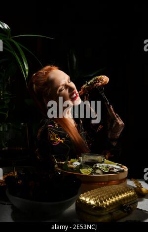 Una ricca donna dai capelli rossi che mangia nel ristorante di pesce tiene un gamberetto a forcella o una langoutine. Tavolo da pranzo al tavolo che serve ostriche fresche Foto Stock