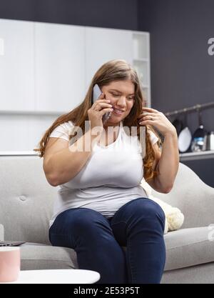 Corpo allegro positivo giovane ragazza che parla al telefono con gli amici mentre si siede a casa. Donna giovane in sovrappeso felice di parlare con nutrizionista. Auto Foto Stock