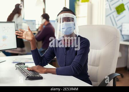 POV dell'imprenditore africano che sventolava in videochiamata con la gente d'affari che svezzava la maschera contro il covid19. Donna che parla con il team durante la conferenza online mentre i colleghi lavorano in background. Foto Stock