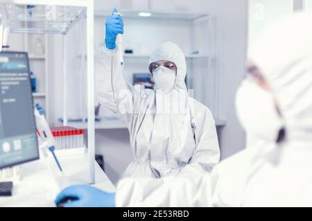 Utilizzo della micropipetta utilizzando un tuta di protezione come precauzione di sicurezza per il coroanvirus. Gruppo di microbiologi in laboratorio di ricerca che conduce esperimenti durante la pandemia globale con covid19. Foto Stock