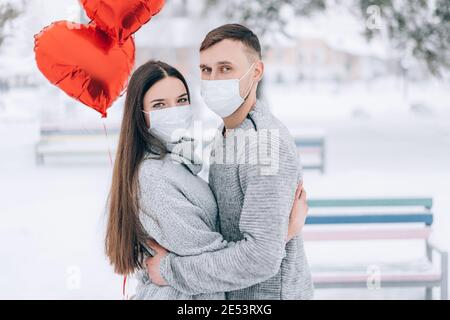 Giovane coppia innamorato nel parco su uno sfondo di neve e palloncini rossi a forma di cuore. Regalo di San Valentino. Maschere mediche sui loro volti. Foto Stock