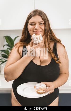 Soddisfatto giovane donna grasso mangiare una torta felice sorridente sulla macchina fotografica che indossa costume da bagno nero. Bella giovane donna chubby mangiare cibo malsano. Ragazza grassa Foto Stock