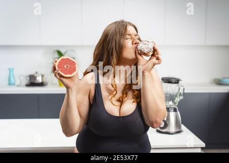 Giovane donna grassa che sceglie che cosa mangiare tenendo una torta e pompelmo. Bella giovane donna chubby mangiare cibo malsano. Ragazza grasso che confronta tra Foto Stock