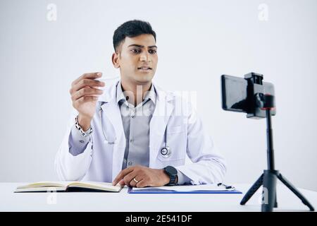 Giovane medico indiano che ha una videoconferenza con il paziente, dando consigli e riempiendo la storia medica Foto Stock