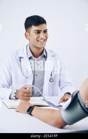 Felice sorridente giovane medico indiano utilizzando tonometro elettronico durante la misurazione pressione sanguigna Foto Stock