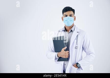 Ritratto di giovane medico serio in maschera medica che tiene cuoio cartella con documenti e guardando la fotocamera Foto Stock