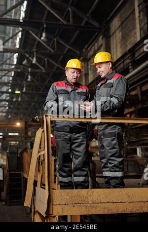 Due lavoratori manuali in abbigliamento da lavoro che utilizzano tablet digitali e discutono lavorare mentre si è in piedi nella pianta Foto Stock