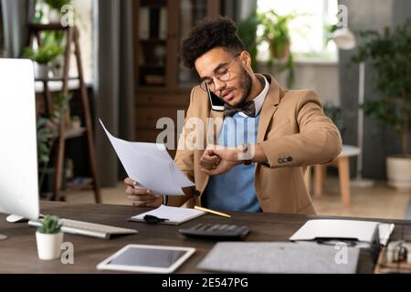 Giovane uomo d'affari occupato seduto sul suo posto di lavoro che lavora con documenti e parlare sul telefono cellulare durante la sua giornata di lavoro a. ufficio Foto Stock