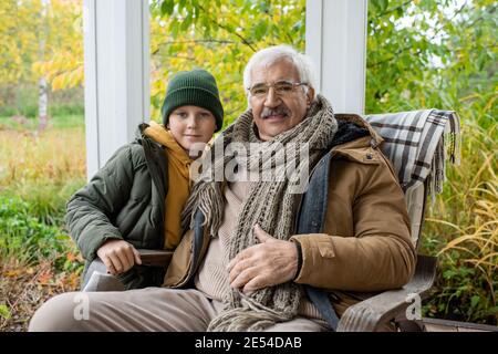 Carino scolaro in bania e giacca calda e suo nonno in sedia dondolo che guarda mentre distendersi contro naturale ambiente Foto Stock