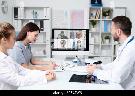 Gruppo di giovani professionisti che si consultano con i colleghi durante la comunicazione online mentre si è seduti davanti al monitor del computer in un ufficio medico Foto Stock