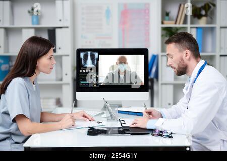 Due giovani professionisti che consultano con il collega durante la comunicazione in linea mentre seduto davanti al monitor del computer in un ufficio medico Foto Stock