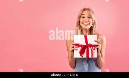 Splendido modello giovane biondo che tiene scatola regalo su sfondo rosa studio, panorama con spazio libero Foto Stock