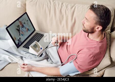 Giovane malato con tonometro che misura la sua pressione sanguigna mentre seduto sul divano sotto la coperta e ascoltando i consigli di medico online Foto Stock