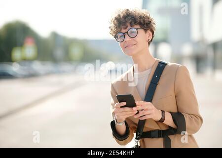 Giovane donna d'affari curly con smartphone in piedi da strada, scorrimento in smartphone e alla ricerca di taxi contro l'ambiente urbano Foto Stock