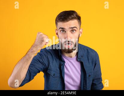 Ritratto di giovane arrabbiato che scuoteva pugno aggraffato alla macchina fotografica e gridava, mostrando la sua protesta su sfondo arancione Foto Stock