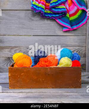 Stuoie a righe colorate e palle di filo di lana brillante in una scatola di legno sullo sfondo della vecchia parete di legno. Foto Stock