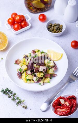 Insalata calda con polpo, patate, pomodori, capperi e limone su un piatto bianco. Primo piano, sfondo bianco. Foto Stock