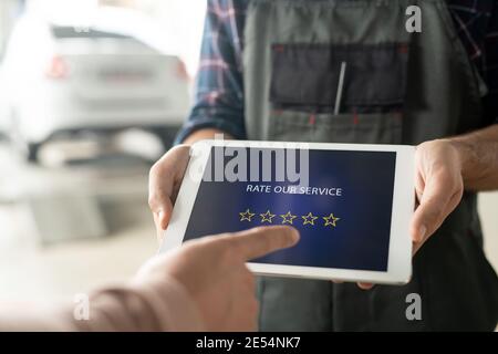 Mani del lavoratore maschile di manutenzione centro auto che tiene il touchpad con cinque stelle sul suo schermo mentre il cliente va a. valuta il servizio Foto Stock