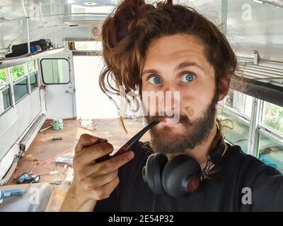 Primo piano ritratto di un giovane dall'aspetto divertente con capelli lunghi, grandi cuffie e un tubo in legno d'epoca all'interno di un autobus in fase di ristrutturazione. Foto Stock