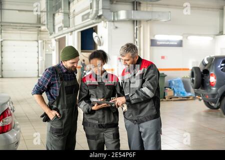 Tre operatori interculturali del centro di manutenzione auto in posizione uniforme in officina tra automobili e discutere di informazioni tecniche Foto Stock