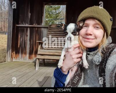 Primo piano ritratto di una giovane donna con lunghi capelli biondi, abiti invernali e un cappello di woo che tiene un po 'nero e bianco chihuahua al viso. Foto Stock