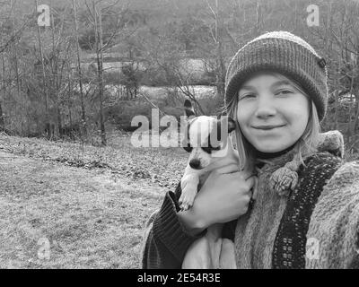 Ritratto bianco e nero di una giovane donna con lunghi capelli biondi, abiti invernali e un cappello di woo che tiene un po 'di chihuahua bianco e nero al viso. Foto Stock