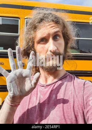 Primo piano ritratto di un giovane dall'aspetto divertente con capelli lunghi, che mostra una mano sporca spolverata accanto a un autobus in fase di ristrutturazione. Foto Stock