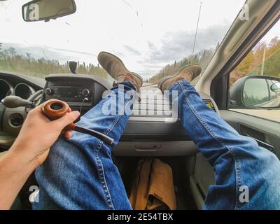 Foto POV dei piedi e delle gambe del fotografo poggiati sul cruscotto dell'auto che attraversa una strada secondaria in campagna. Foto Stock