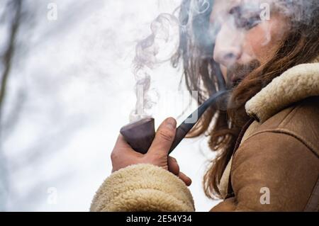 Ritratto di un giovane uomo con un cappotto invernale in pelle di pecora che fuma un tubo all'aperto con grandi spruzzi di fumo bianco di fronte al viso. Foto Stock