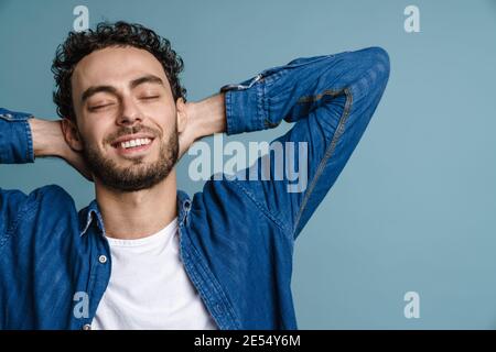 Felice bel ragazzo che posa con le mani aggrappate dietro il collo isolato su sfondo blu Foto Stock