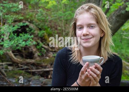 Ritratto di una bella giovane ragazza seduta su una roccia sulla riva di un fiume forestale, tenendo una tazza da tè cinese con entrambe le mani e sorridendo alla macchina fotografica. Foto Stock