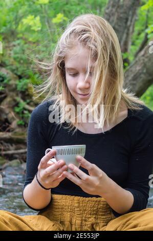 Ritratto di una giovane ragazza graziosa seduta su una roccia sulla riva di un fiume di foresta, tenendo una tazza di tè cinese con entrambe le mani e guardandola giù. Foto Stock