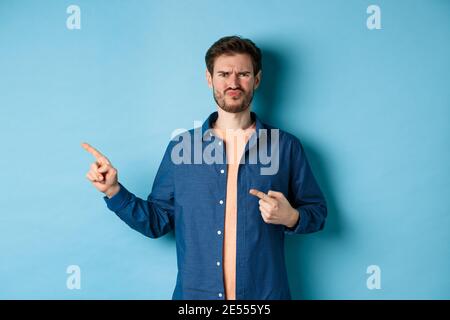 Deluso e scettico ragazzo accigliato, labbra pucker e dita di puntamento sinistra a spazio vuoto, lamentando di cattivo cosa, in piedi sconvolto su blu Foto Stock