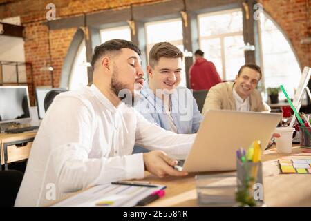 Discutere, parlare. Giovani colleghi caucasici che lavorano insieme in un ufficio utilizzando dispositivi e gadget moderni. Guarda occupato, attento, allegro, riuscito. Concetto di business, ufficio, finanza. Foto Stock