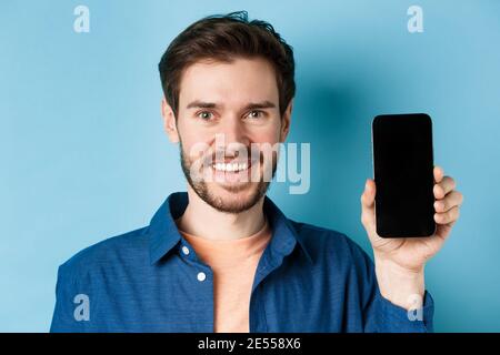 Primo piano di un uomo europeo sorridente che mostra lo schermo vuoto dello smartphone e mostra l'app, in piedi su sfondo blu Foto Stock